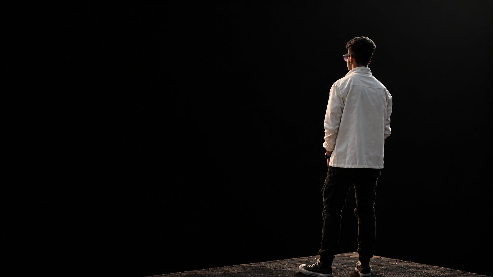 Aswin Paudel seen from behind in a white jacket, standing on a sunlit ledge and looking out into the dark